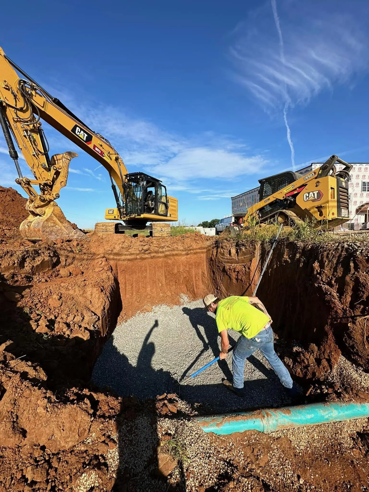 Construction worker working in a deep trench with excavator nearby.