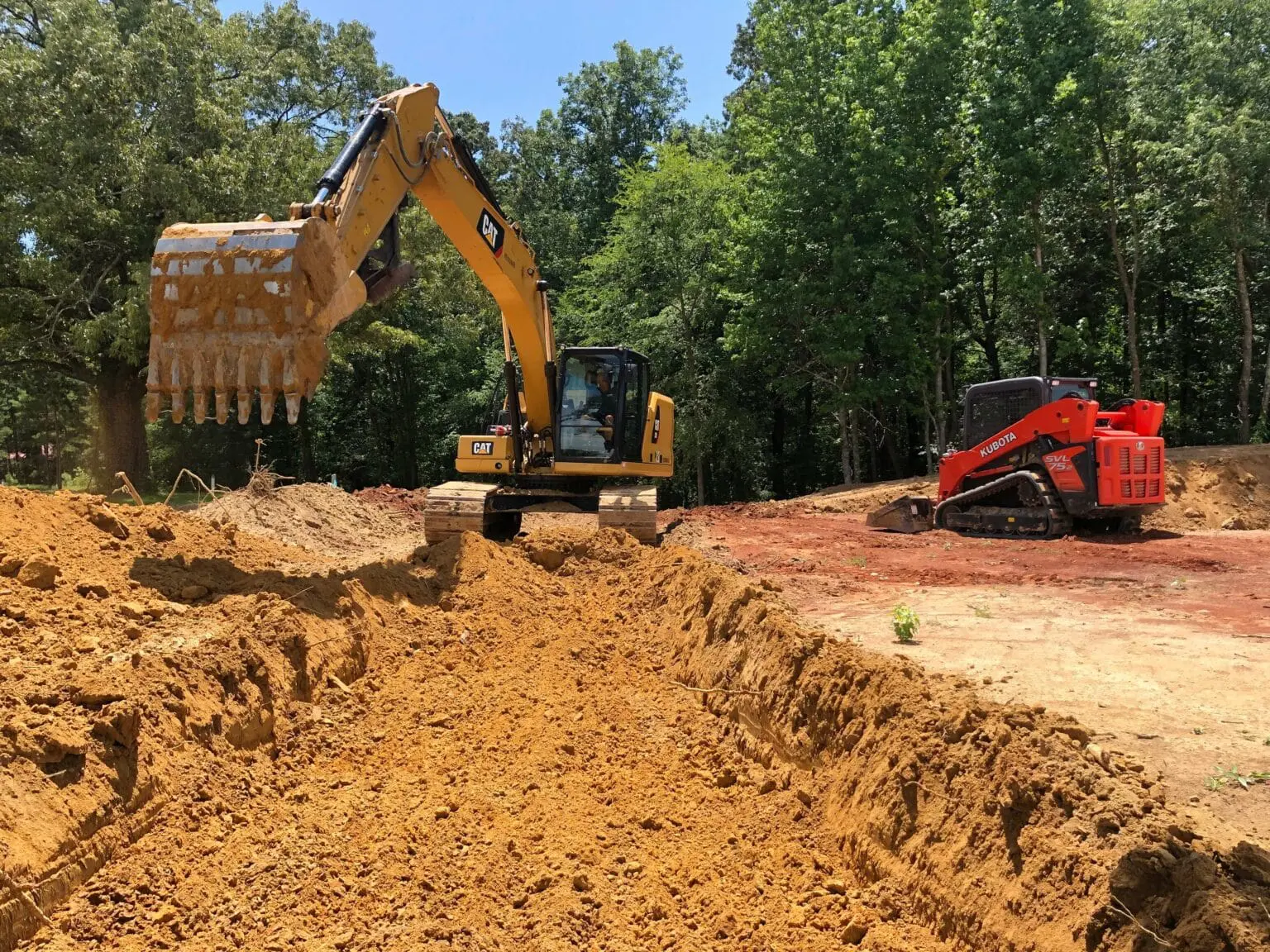 Excavator digging a trench at a construction site surrounded by trees.